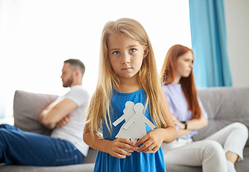 A solemn child holds paper dolls, with parents arguing in the background.